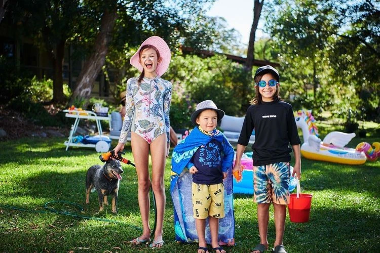 Three children and a dog in a grassy yard, wearing swimsuits and hats, with inflatable toys, trees, and houses in the background on a sunny day.