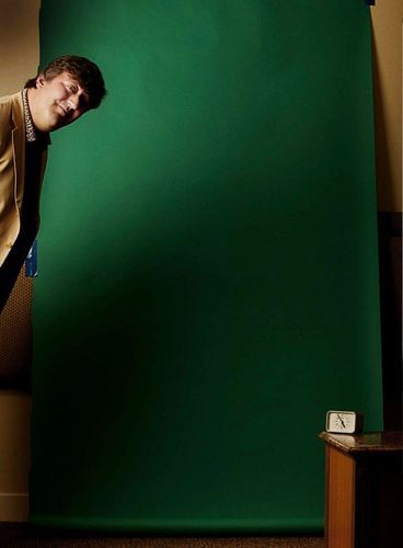 A man in a blazer looks around a large, dark-green seamless background in an indoor setting, with a wooden table and alarm clock.