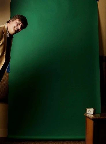 A man in a blazer looks around a large, dark-green seamless background in an indoor setting, with a wooden table and alarm clock.
