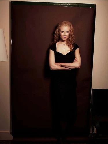 Portrait of fair-skinned woman with arms crossed in black dress in front of backdrop. Red hair, jewelry. Shoulder-length shot.