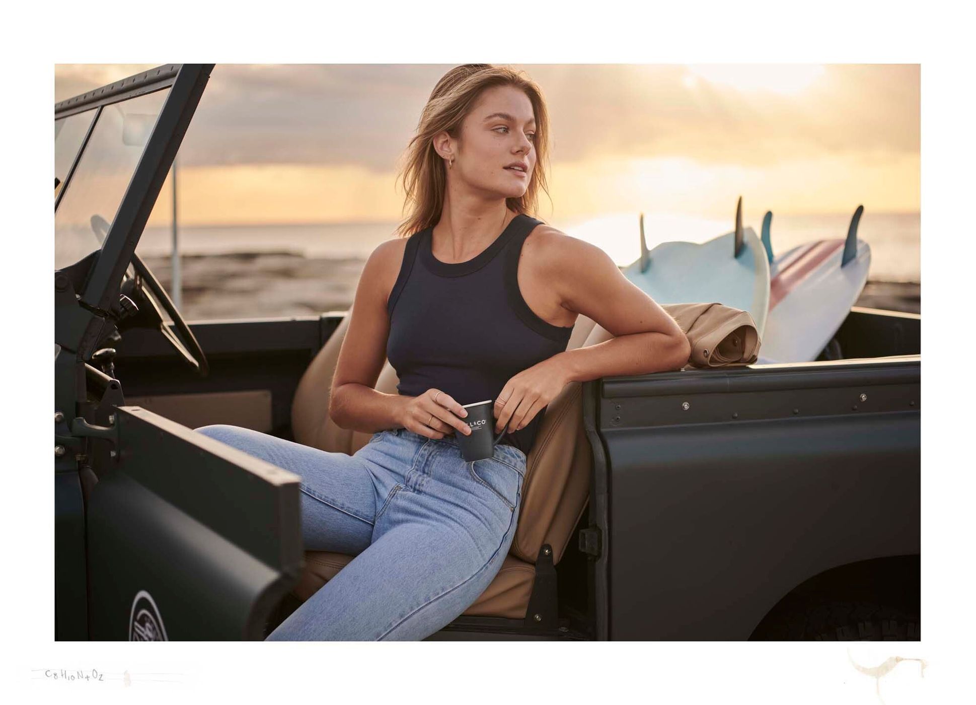 Woman in tank top and jeans sits in olive car with surfboards at beach against sunset. Holding cup, looking to left.