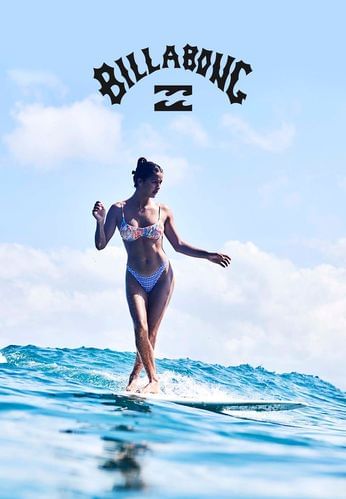 A woman in a bikini surfs on a surfboard in the ocean under a blue sky with the Billabong logo displayed prominently.