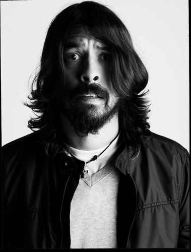 Black and white portrait of a man with long hair and a beard wearing a v-neck sweater and jacket against a white backdrop.