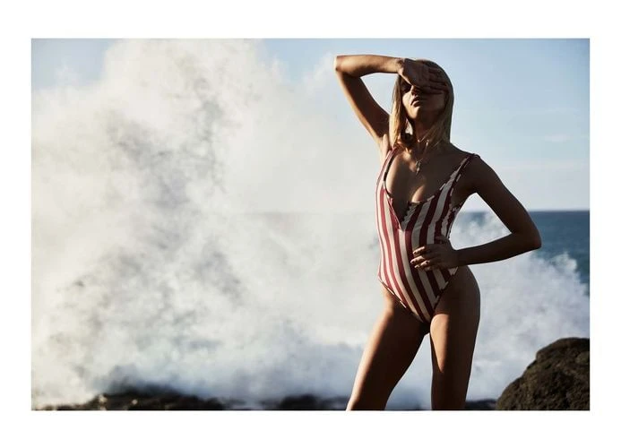 Woman in a striped swimsuit shields her eyes from the sun on a rocky beach with a large wave crashing in the background.