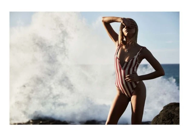 Woman in a striped swimsuit shields her eyes from the sun on a rocky beach with a large wave crashing in the background.