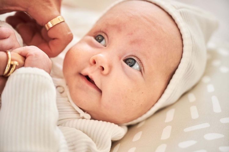 Close-up of a baby lying on a patterned surface, gazing upward, with an adult hand and gold rings visible near the baby's face.