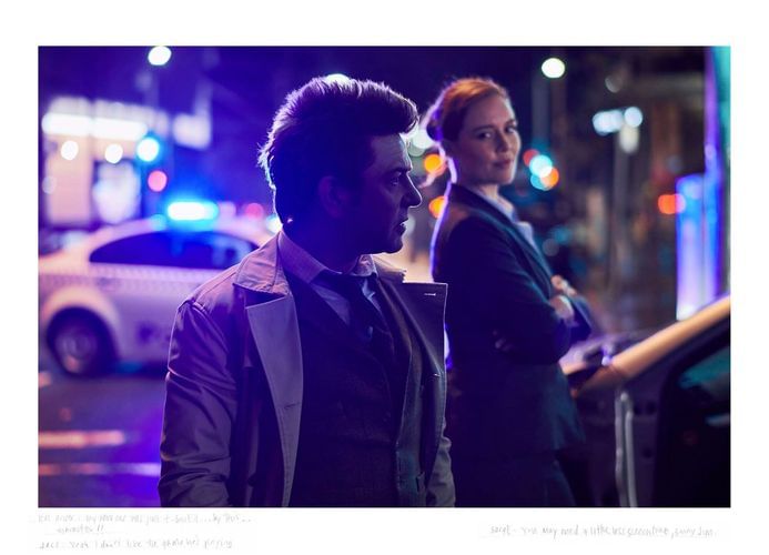 Two adults stand outdoors at night, near a police car with flashing lights, creating an urban street scene with cool lighting.