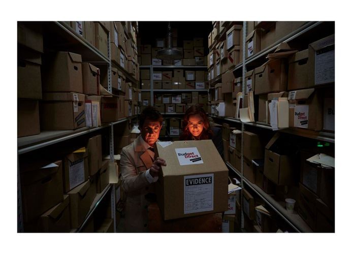 Two investigators in a storage room examine a box labeled 'Evidence,' surrounded by rows of cardboard boxes in dim, film-noir style lighting.