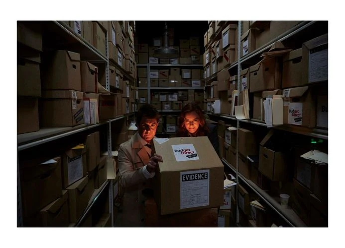 Two investigators in a storage room examine a box labeled 'Evidence,' surrounded by rows of cardboard boxes in dim, film-noir style lighting.