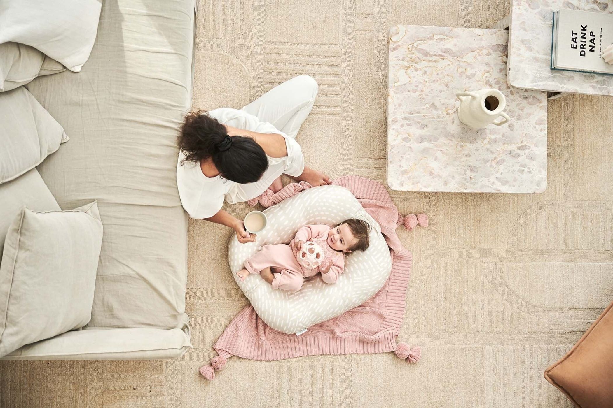 Overhead shot of a woman sitting next to a baby lying in a baby lounger on a rug in a bright, neutral-toned home.