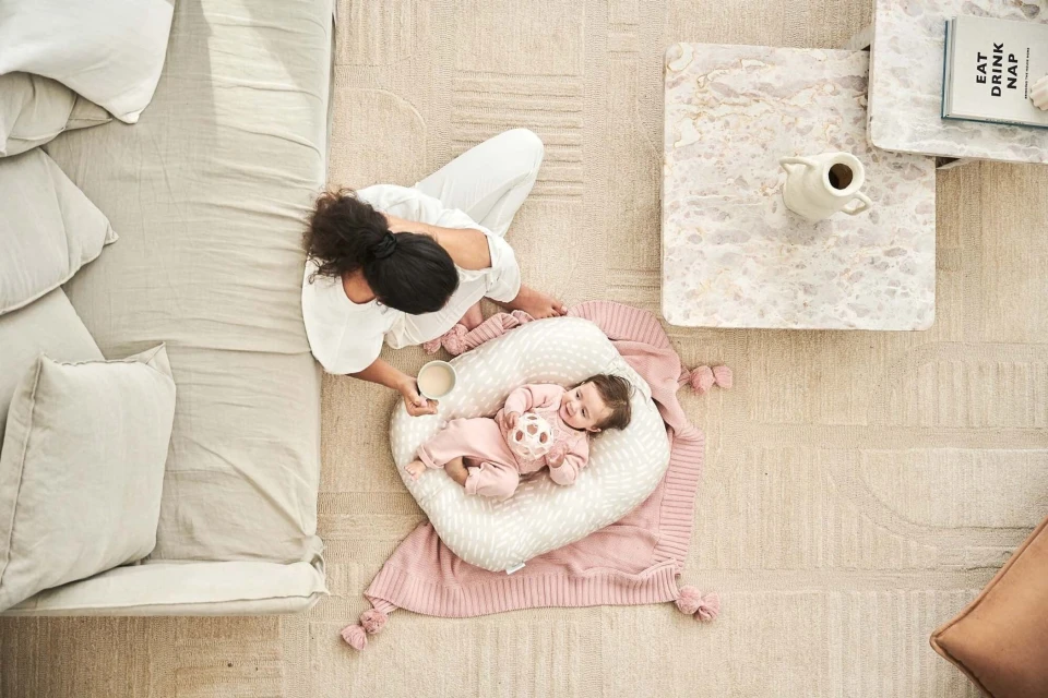 Overhead shot of a woman sitting next to a baby lying in a baby lounger on a rug in a bright, neutral-toned home.