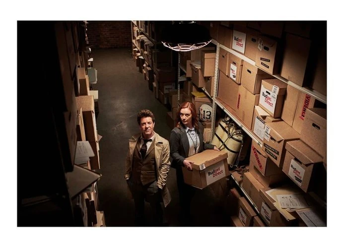 Two people stand in a storage room with cardboard boxes stacked on shelves, the woman holds a 'Budget Direct' branded box.