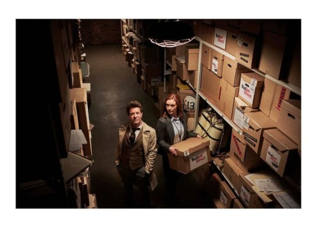 Two people stand in a storage room with cardboard boxes stacked on shelves, the woman holds a 'Budget Direct' branded box.