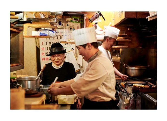 Busy commercial kitchen scene: chefs cooking in uniform, an elderly woman in a black hat, and stainless steel cookware and equipment.