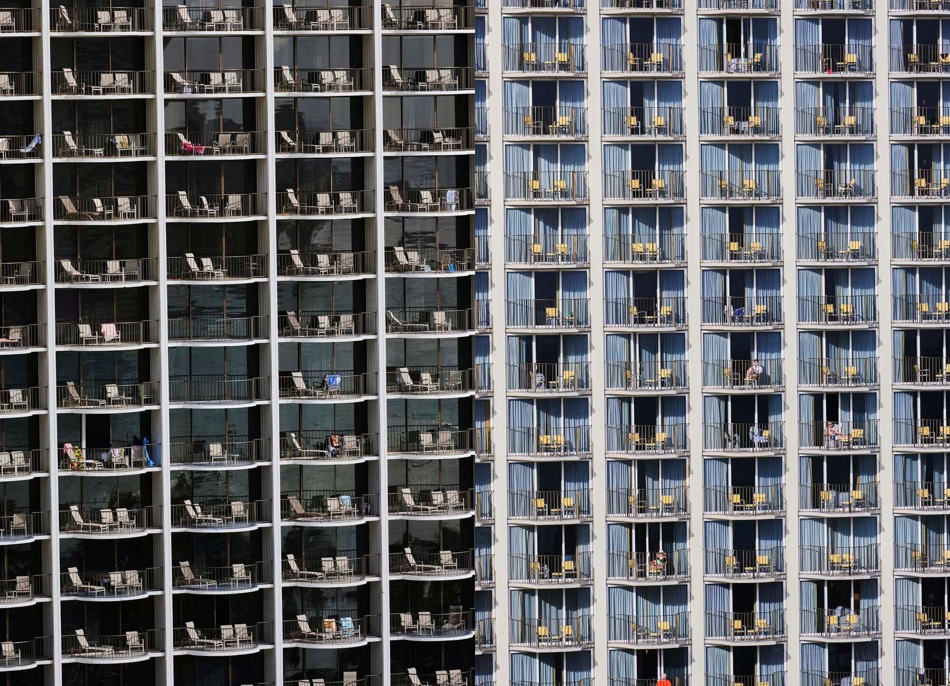 Exterior of a tall hotel building with multiple balconies and chairs, showcasing architectural details and a sunny, vacation-oriented scene.