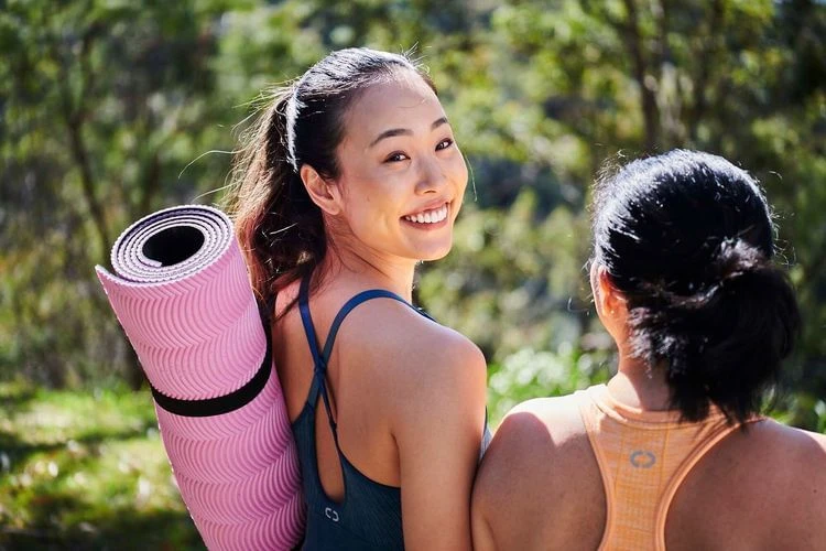 Smiling Asian woman with a pink yoga mat over her shoulder and another woman outdoors in natural sunlight. Fitness and wellbeing theme.