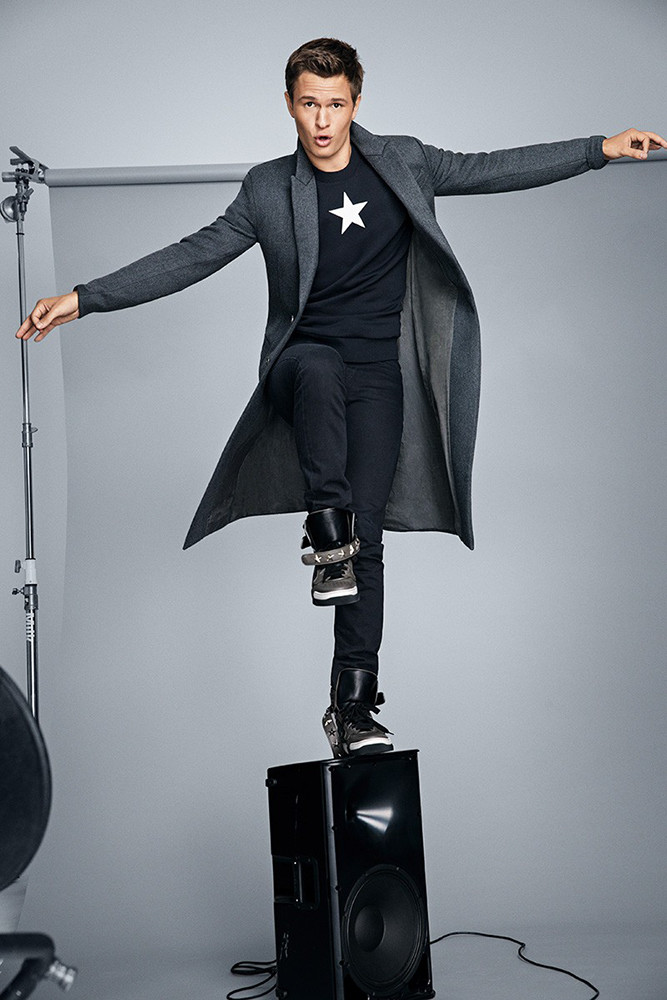 Stylish man in a gray coat poses on a speaker in a photo studio with a white backdrop and professional lighting equipment visible.
