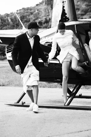 A man and woman exiting a helicopter on a tarmac, the man holding the woman's hand for support in a black and white shot.