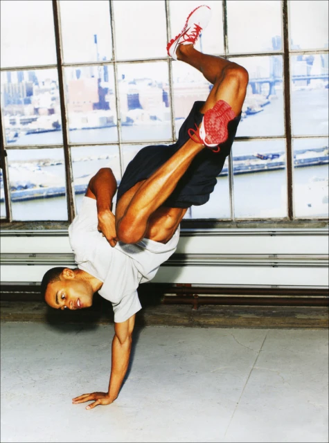 An athletic African-American man performs a handstand breakdance move indoors with urban background, showcasing strength and skill in dance.