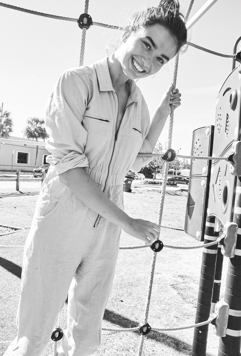 Smiling adult woman in a jumpsuit posing on a playground climbing structure in a park on a sunny day in a black and white photograph.