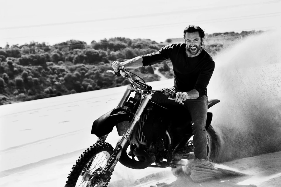 A man rides a dirt bike on a sand dune near the ocean, smiling, wearing a dark shirt and jeans. Trees and the ocean are in the distance.