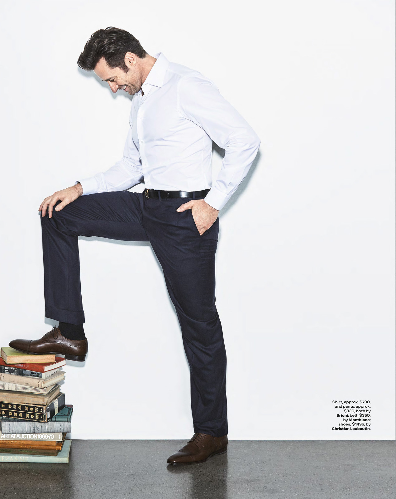 A man in a white shirt and black pants poses with one foot on a stack of books indoors against a white wall with copy space. 