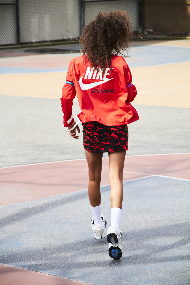 Woman in red Nike jacket and skirt walking on a colourful concrete court, seen from the back. Stylish athletic apparel and outdoor setting.