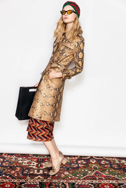 A stylish woman poses in a snake-print jacket, culottes, turban, and sunglasses against a white background and patterned vintage carpet.