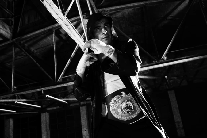 Black and white portrait of a male boxer in a hooded robe wearing a championship belt and taped hands in a warehouse