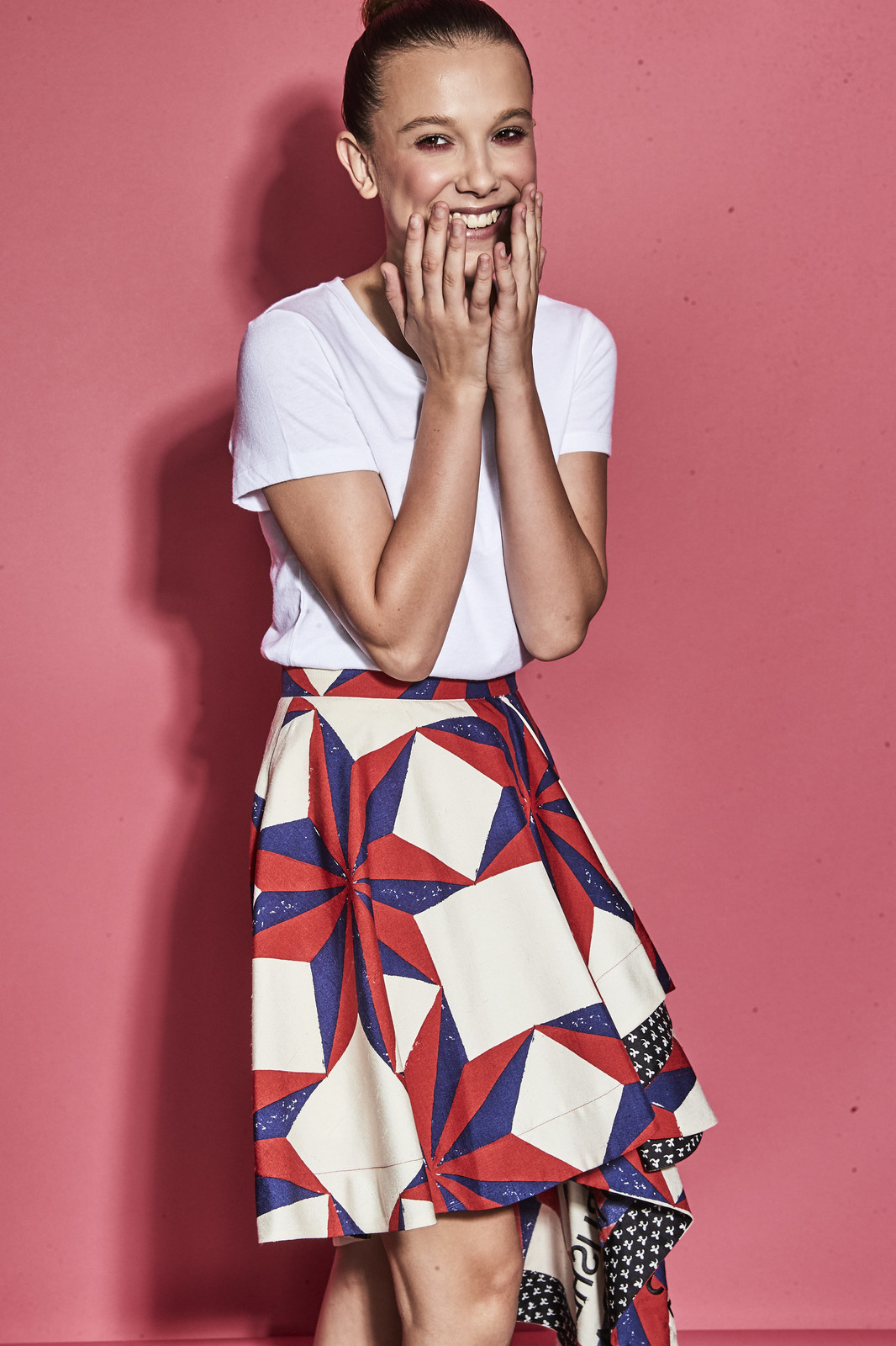 Millie Bobby Brown smiles with hands near face, wearing a white t-shirt and patterned skirt against a pink backdrop in a studio shot.