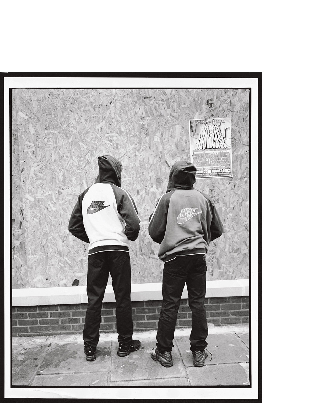 Two hooded figures wearing Nike jackets stand against a boarded wall with a poster, captured in a black and white editorial photograph.