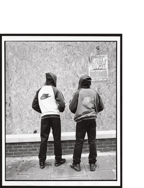 Two hooded figures wearing Nike jackets stand against a boarded wall with a poster, captured in a black and white editorial photograph.
