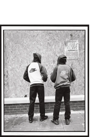 Two hooded figures wearing Nike jackets stand against a boarded wall with a poster, captured in a black and white editorial photograph.