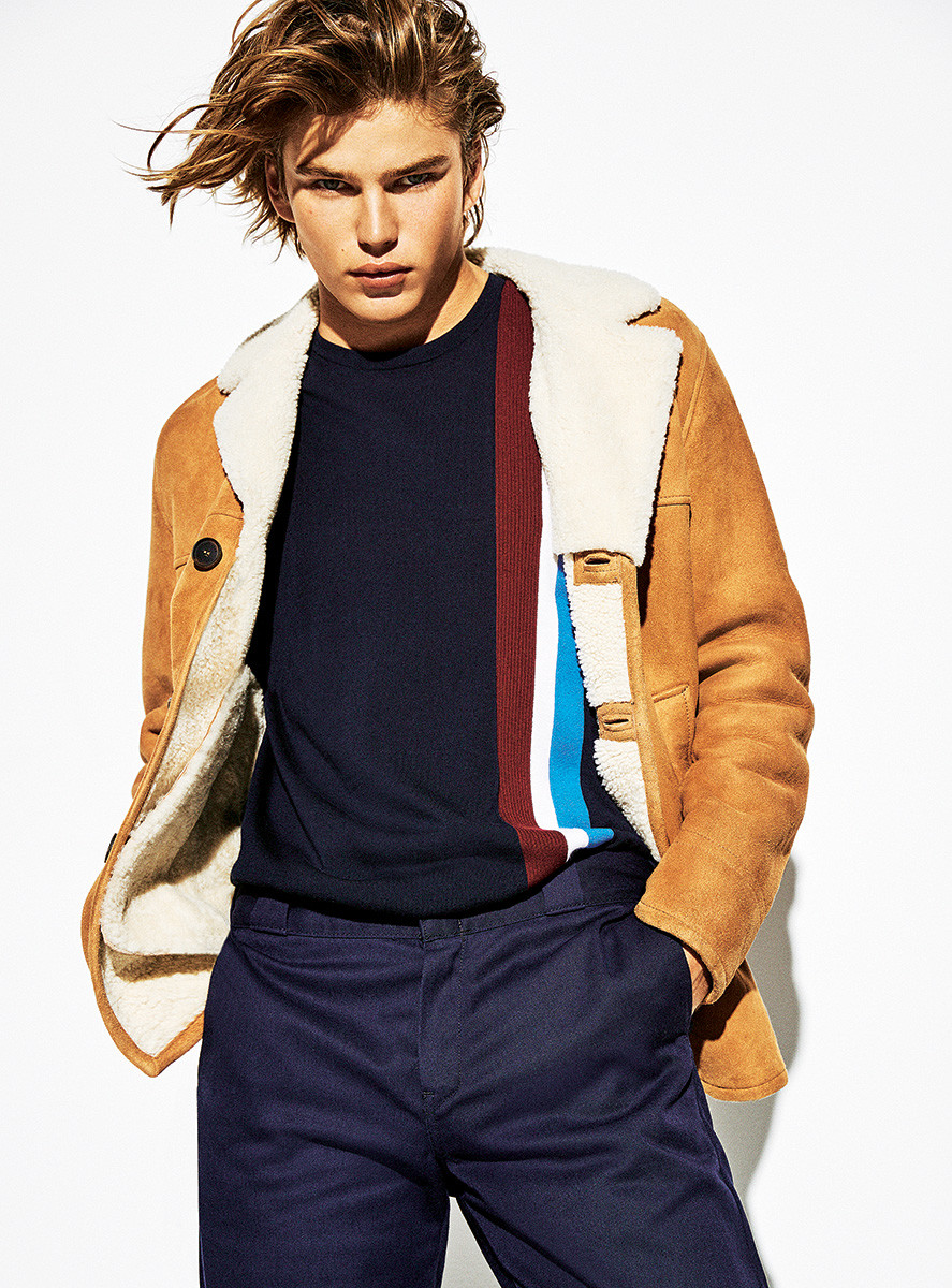 Blond male fashion model poses in a tan shearling coat and dark pants against a white background in a studio shot, showcasing modern style.