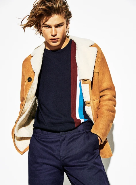 Blond male fashion model poses in a tan shearling coat and dark pants against a white background in a studio shot, showcasing modern style.