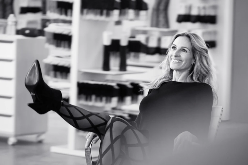A smiling woman in patterned tights sits in a chair with her feet up, showcasing black high heels and a confident pose in a monochrome shot.