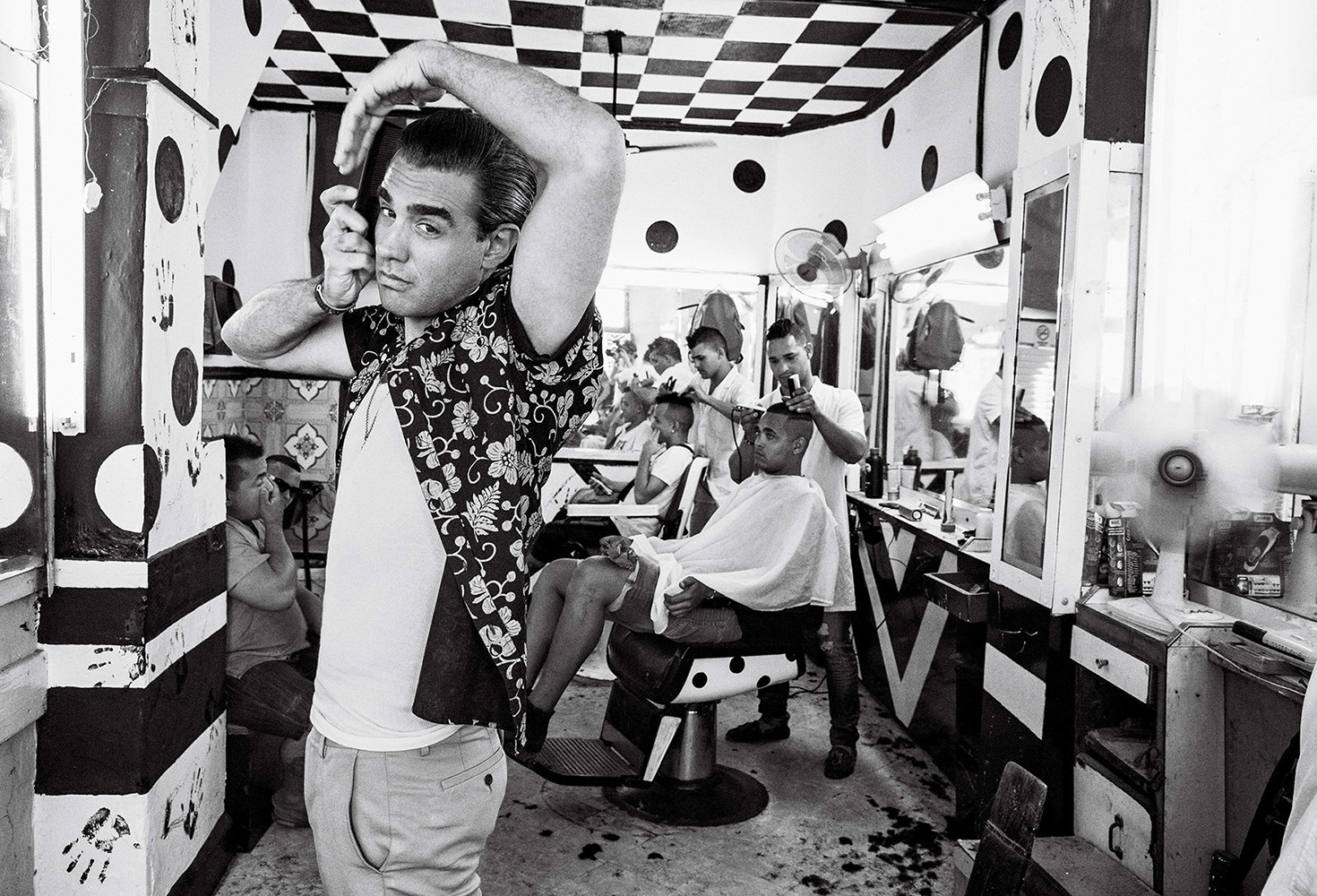 Black and white shot in a barbershop showing a man styling his hair while other barbers give haircuts in the background.
