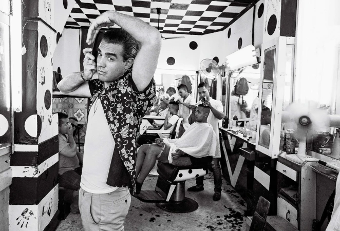 Black and white shot in a barbershop showing a man styling his hair while other barbers give haircuts in the background.