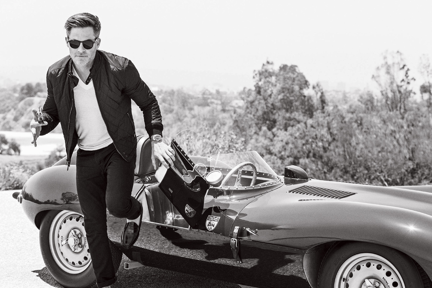 Black and white shot of a man exiting a vintage car with sunglasses, jacket, and loafers in an outdoor, tree-lined setting.