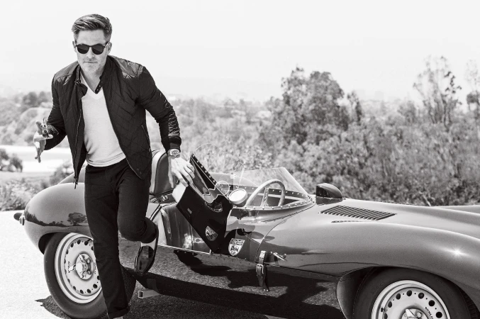 Black and white shot of a man exiting a vintage car with sunglasses, jacket, and loafers in an outdoor, tree-lined setting.