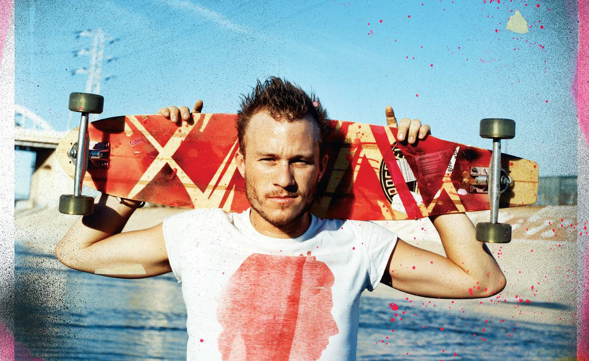 Portrait of actor Heath Ledger holding a red skateboard across his shoulders against a bright sky background.
