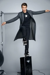 Ansel Elgort balancing playfully on a black audio speaker against a grey studio backdrop, wearing a dark coat and star-print sweater.