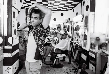 Monochrome portrait of a man in a patterned short-sleeve shirt posing in a barber shop by Ben Watts