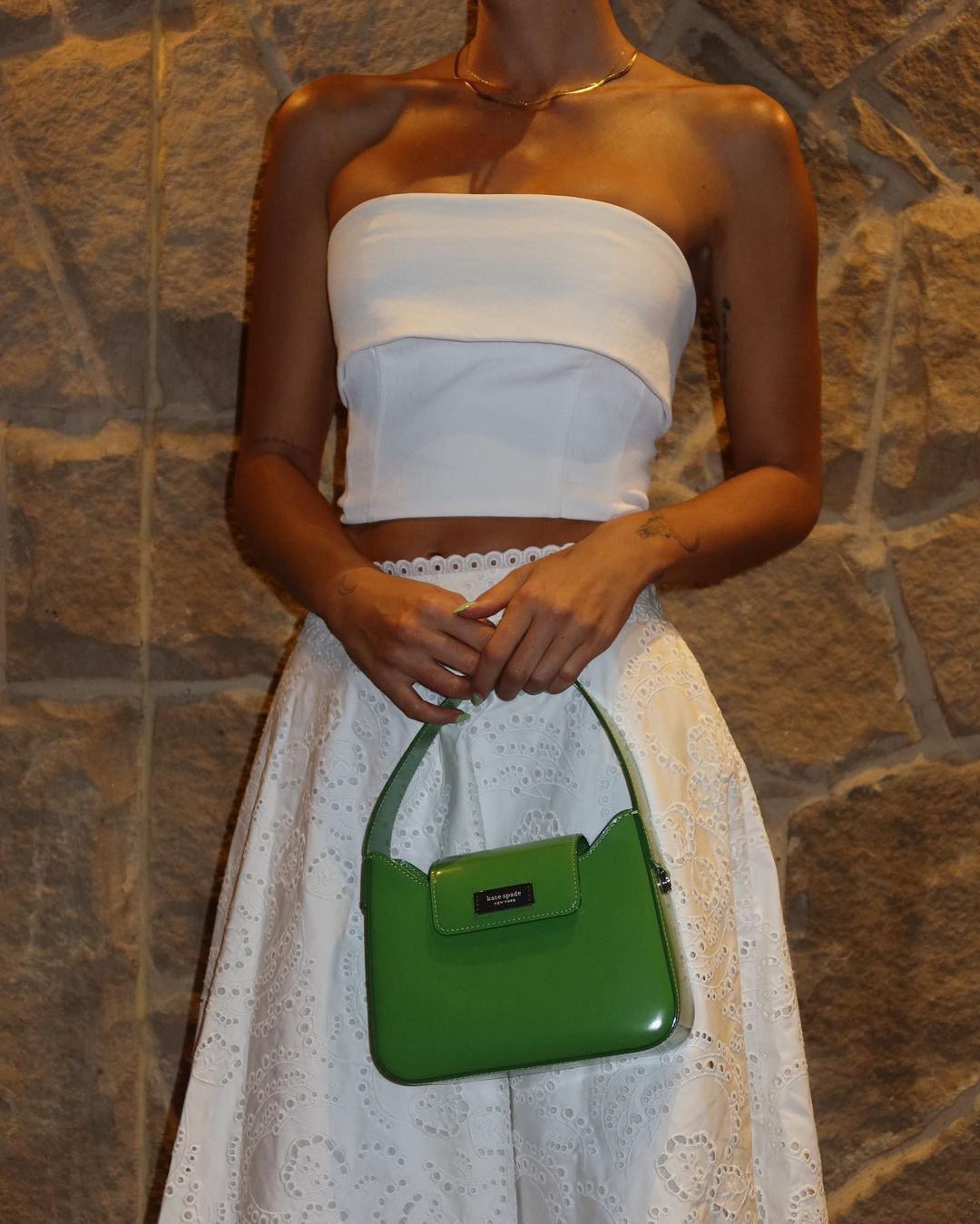 Fashionable woman in white strapless top and eyelet skirt holding green Kate Spade purse in front of tan stone wall.