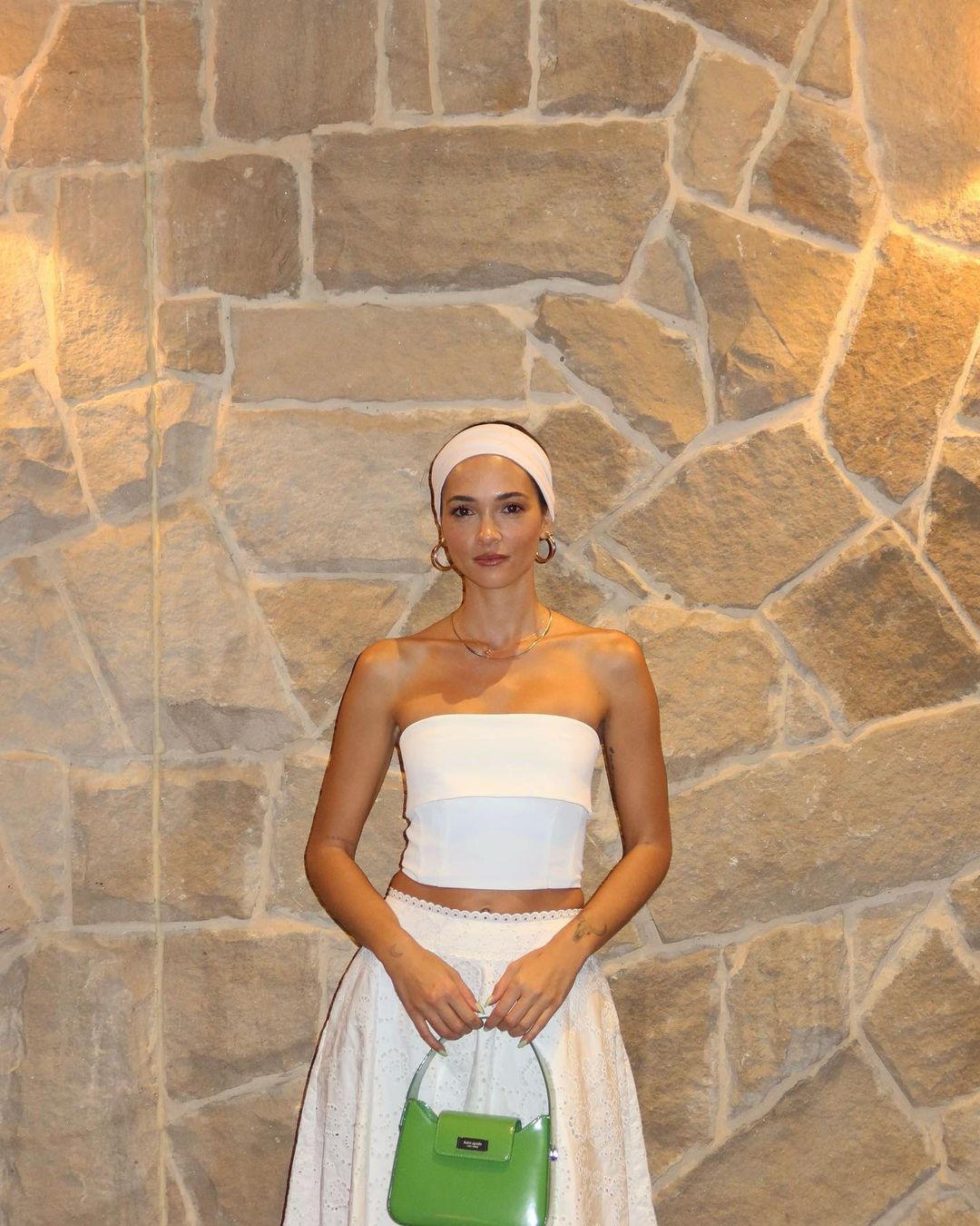 A woman with a headband and green purse poses in front of a stone wall, wearing a white top and skirt, accessorized with earrings and a necklace.