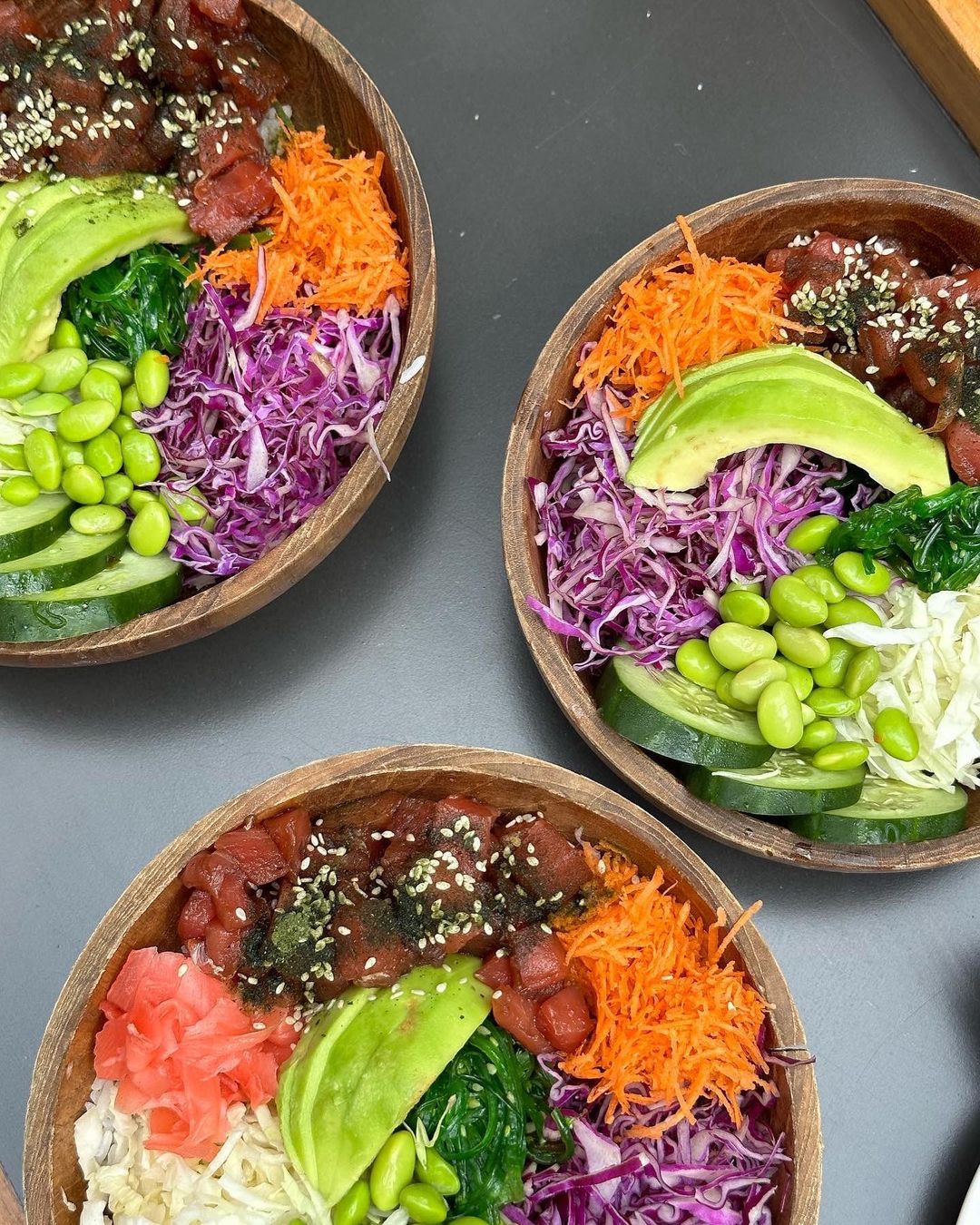 Three wooden bowls are filled with poke salad including tuna, carrots, avocado, seaweed, cabbage, and rice on a gray surface.
