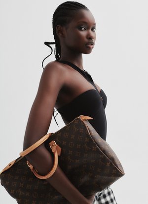 Portrait of a young woman with braided hair, wearing a black top and holding a brown monogrammed handbag against a white backdrop.