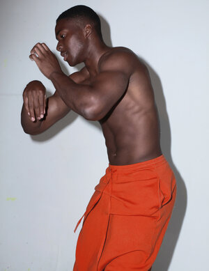 Shirtless athletic man in orange pants poses against a plain wall in a studio setting, showing muscular physique and toned torso.