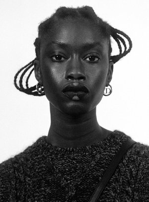Monochrome portrait of a dark-skinned woman with braided hair, wearing a knit sweater and hoop earrings against a white backdrop.