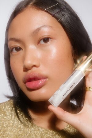 Close-up studio shot of a person holding MECCA MAX lip gloss, showcasing glossy lips, hair clips, and a shiny gold top against a neutral background.
